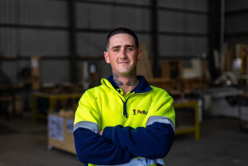 Close up shot of a ReBuild employee standing with arms folded, smiling in front of a workshop