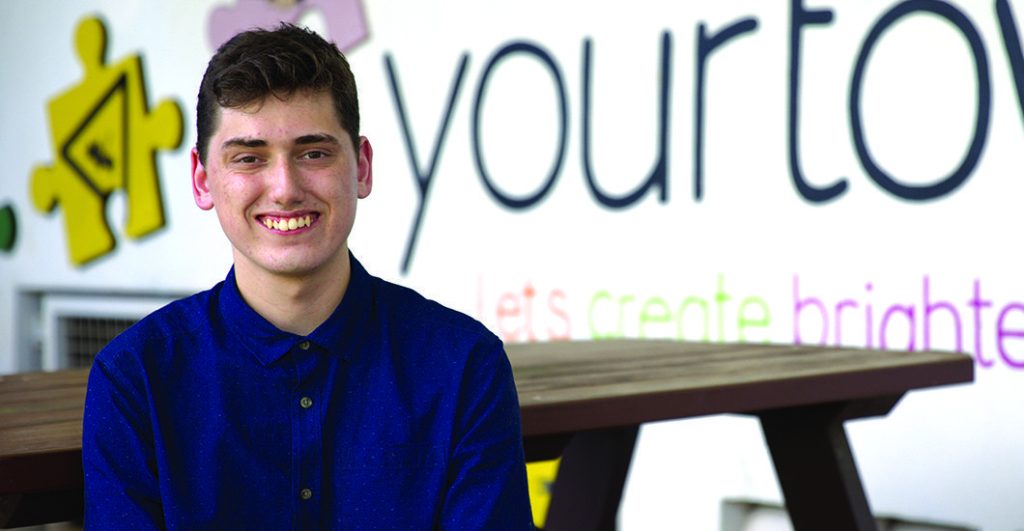 Cropped image of a young man sitting at a wooden table smiling in front of a wall featuring a slogan