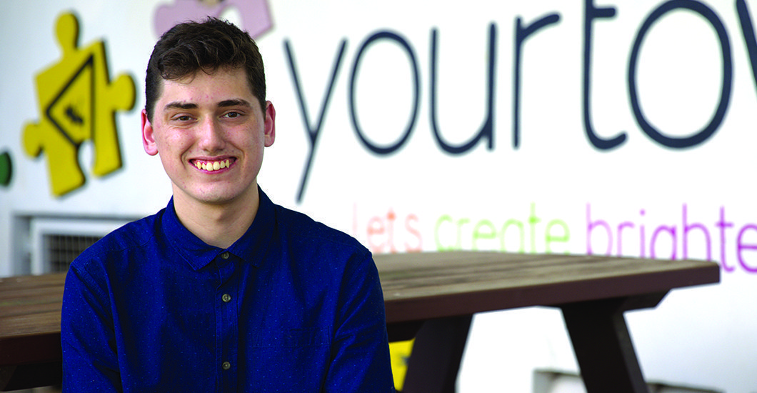 Cropped image of a young man sitting at a wooden table smiling in front of a wall featuring a slogan