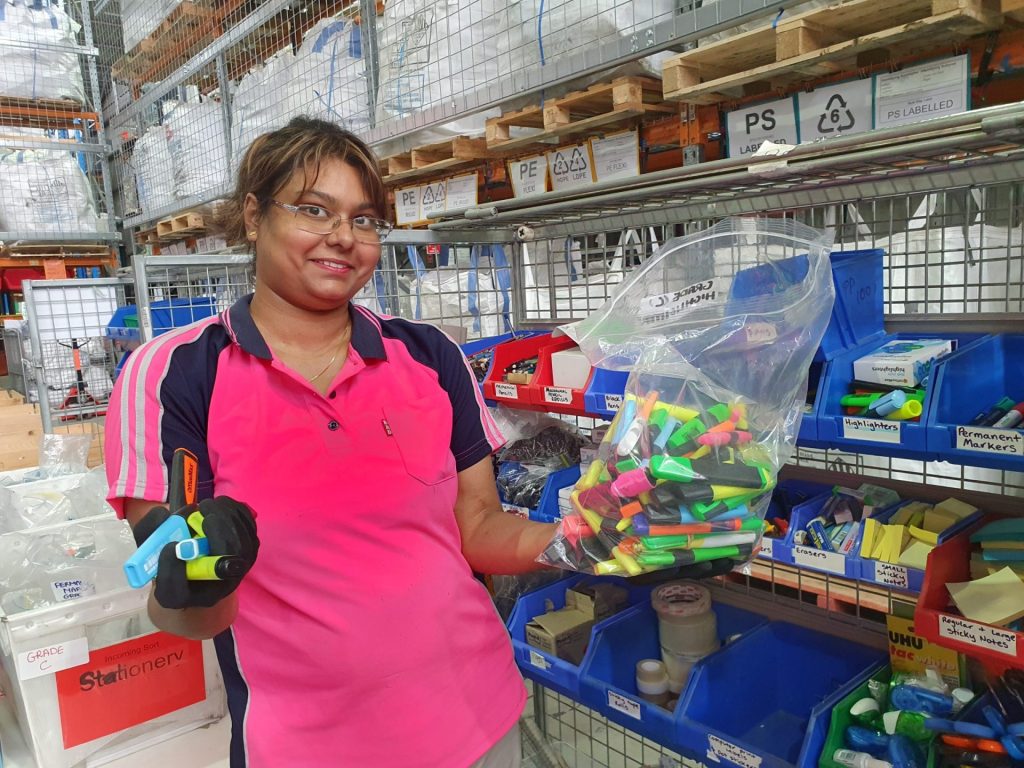 A woman in bright pink polo shirt collects stationary in a plastic bag in a warehouse