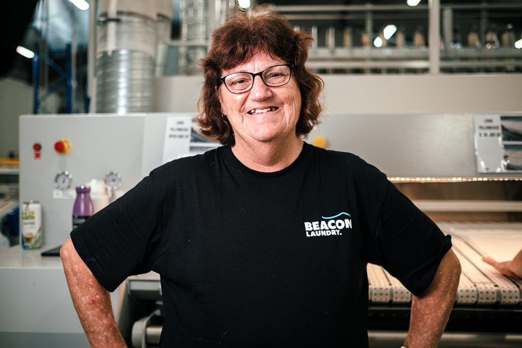 Leigh, a Beacon Laundry employee, standing in front of laundry machinery smiling.